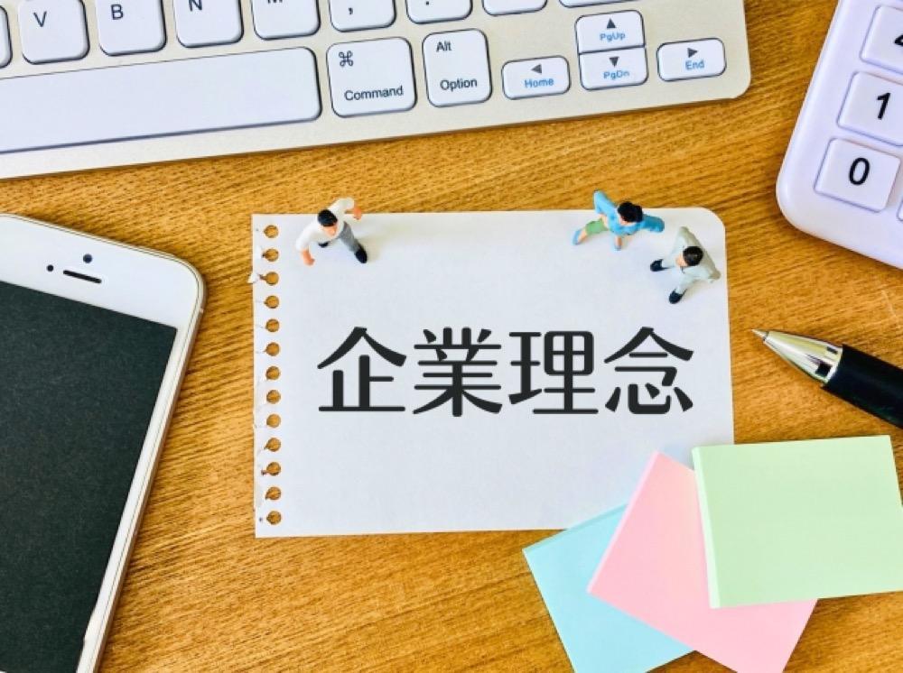 「企業理念」と書かれた紙とミニチュアの人物、パソコン周辺機器が並ぶデスク