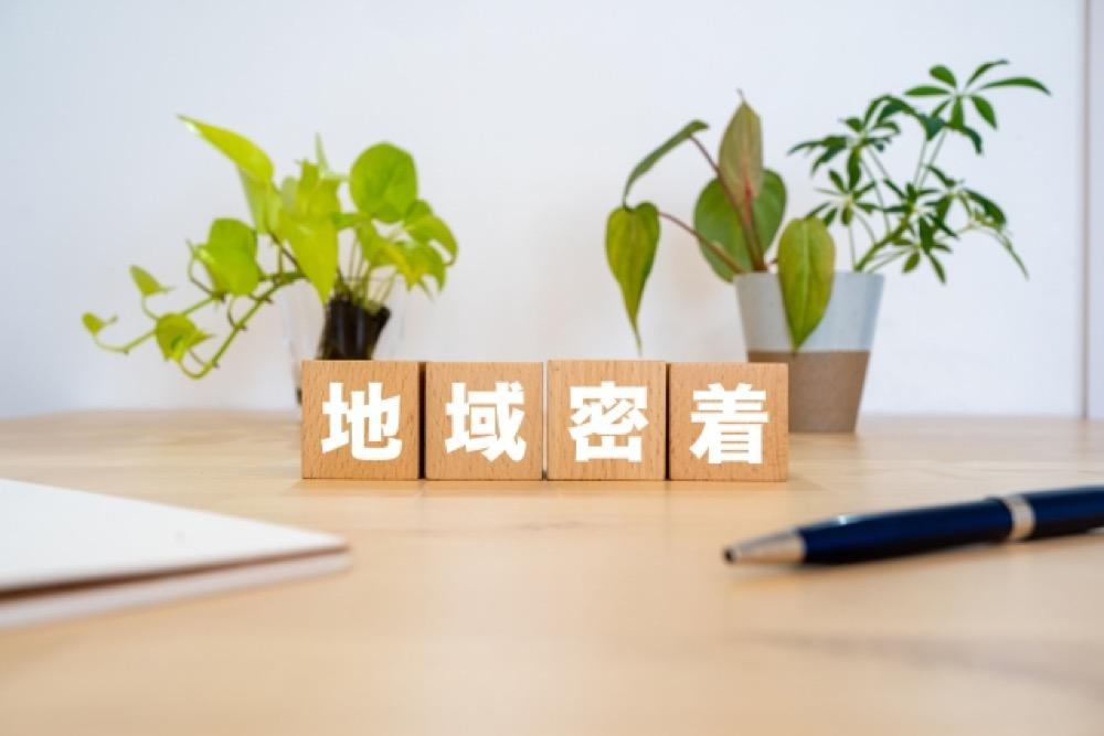 木製ブロックに「地域密着」と書かれた文字、背景には観葉植物とペン