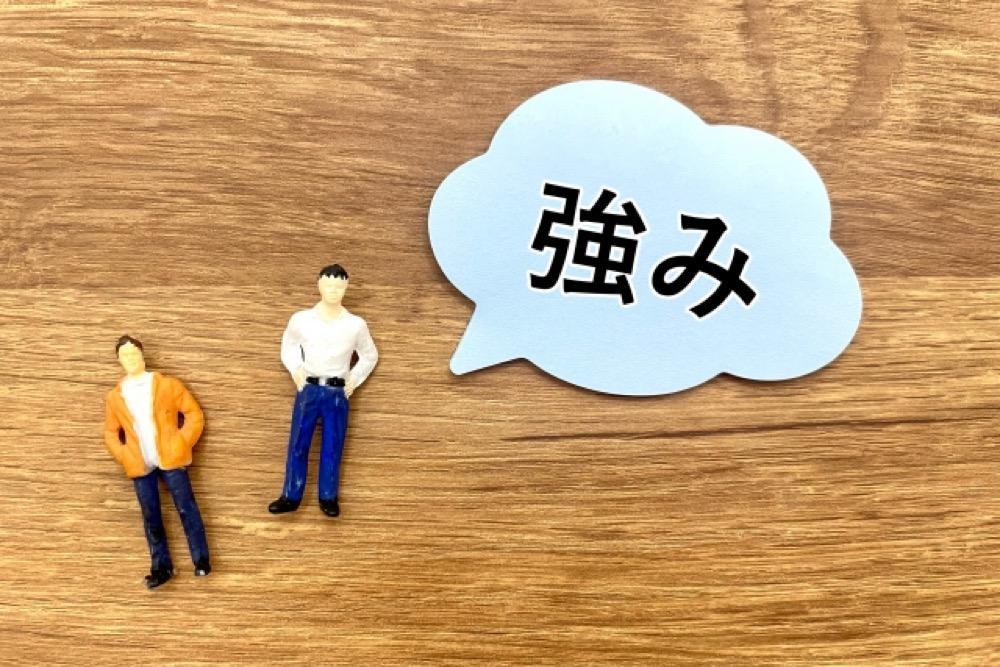 木目の背景に「強み」と書かれた吹き出しと2人の男性フィギュア