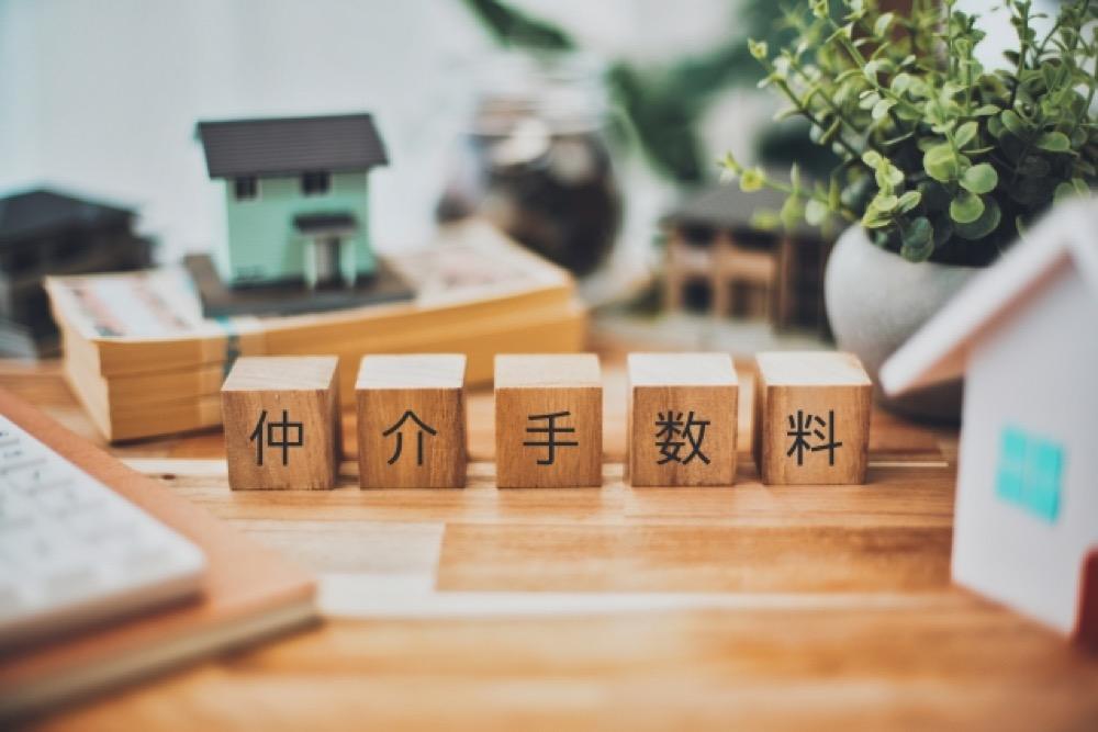 「仲介手数料」と書かれた木製ブロックと住宅模型、観葉植物が並ぶテーブル